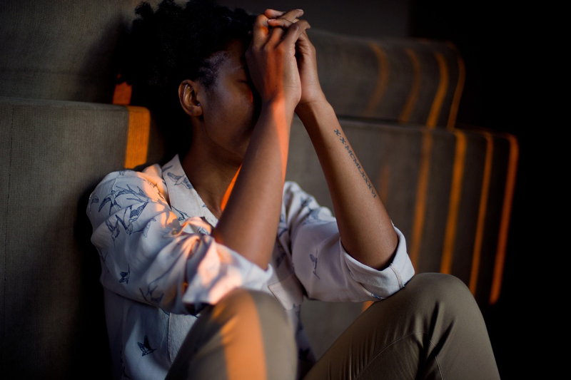 An image of a person leaning against a wall with their elbows resting on their knees and their hands clasped in front of their face. 